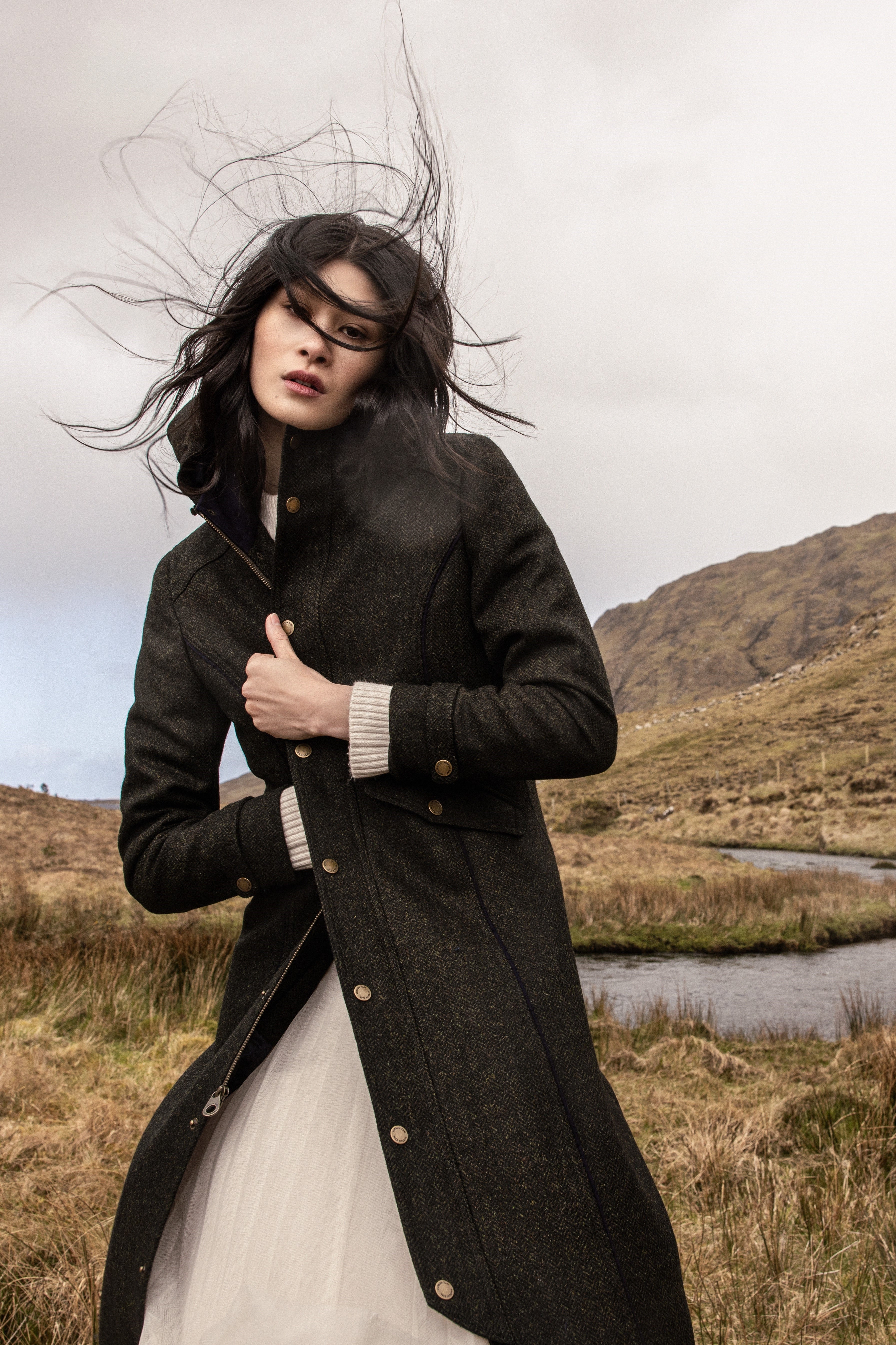 Female model standing in Irish countryside with river behind her wearing a green tweed coat and her hair being blown