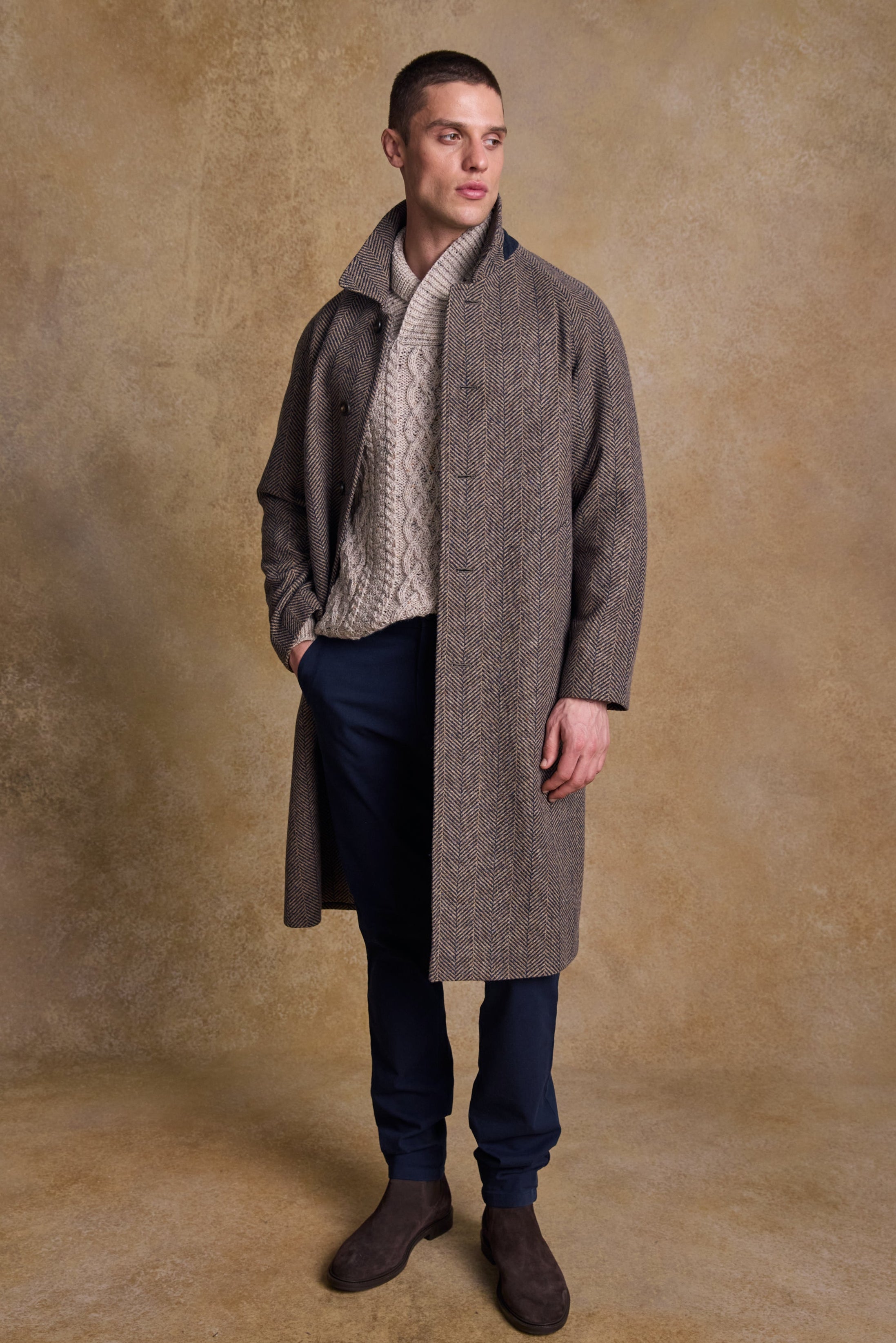 Man wearing a long donegal tweed coat over a sweater and jeans against a textured beige background