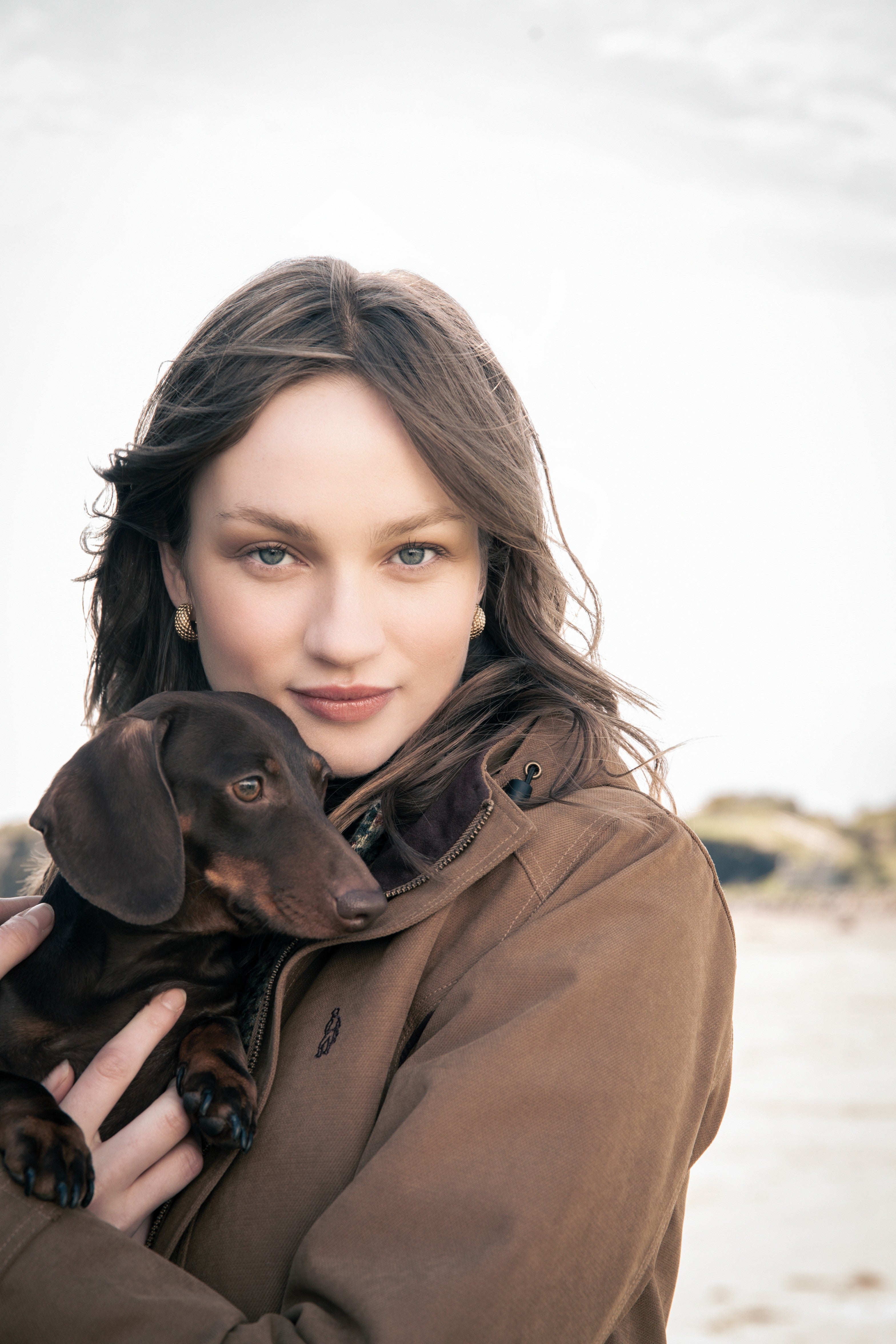 Woman on beach holding sausage dog in Una waterproof jacket camel.