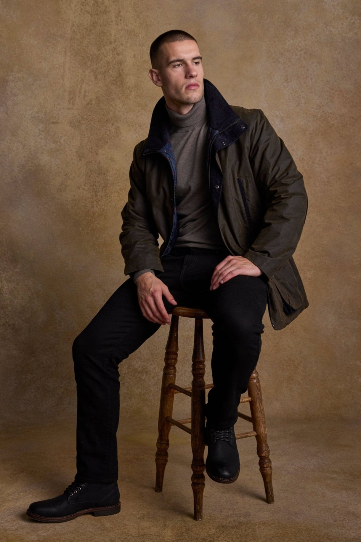 Man sitting on a wooden stool against a textured brown background with a wax jacket on 