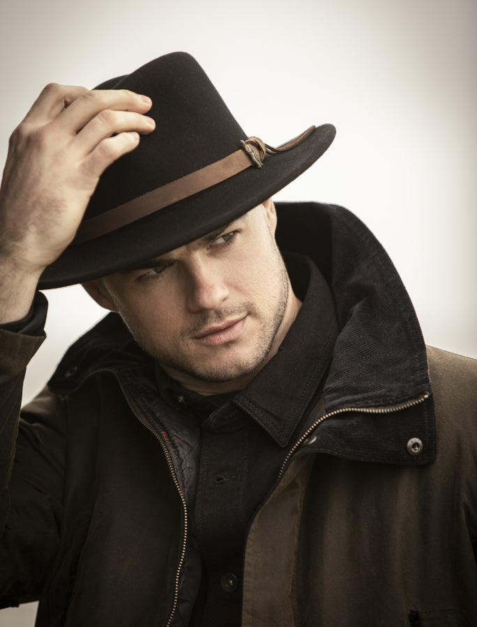 Man taking off wide brimmed Boston felt hat in black while wearing a wax jacket